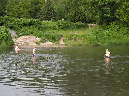 Three shirtless people, standing in a river with water up to their waists, with a grassy bank and trees behind them